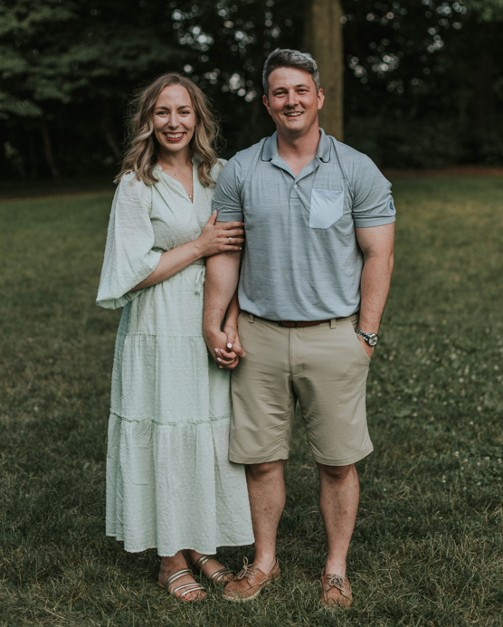Lauren C on the left and her husband on the right are standing next to each other holding hands in a field of grass